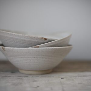 A stack of pale coloured ceramic pasta bowls, shown on a rustic wooden table top against a light grey background. 