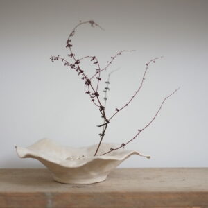 A shallow fluted vase made from an off-white stoneware. Shown on a wooden shelf against a white backdrop. There is a single stemp of bracken displayed in the vase. 