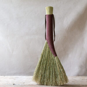 light brown hand broom bound with brown cord. Shown standing on a light wooden table against a linen background