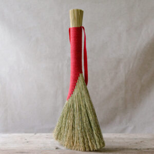 Light brown handbroom bound in red cord and shown on a pale wooden board against a light linen background. 