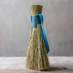 natural fibre hand brush, bound with a teal blue cord. Show standing on a pale wooded table against a light fabric backdrop. 
