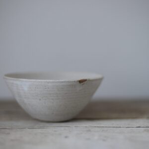 A cone shaped stoneware bowl with a  pale matt finish, shown on a rustic wooden tabletop against a light grey background.
