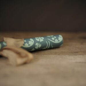 A bluey green coloured wrapping paper covered in a repeat wildflower pattern. Photographed rolled and tied up in a bow with a light brownish pink, silk ribbon. Laying on a rustic wooden tabletop against a very dark background.