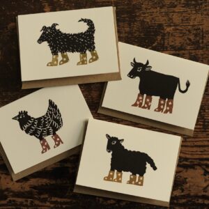 Set of four card designs shown on a wooden table. Designs include a lino cut print of a dog, cow, chicken and sheep wearing cowboy boots. 
