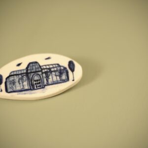 A white and blue ceramic brooch with a naive illustration of the a botanical glass house.  